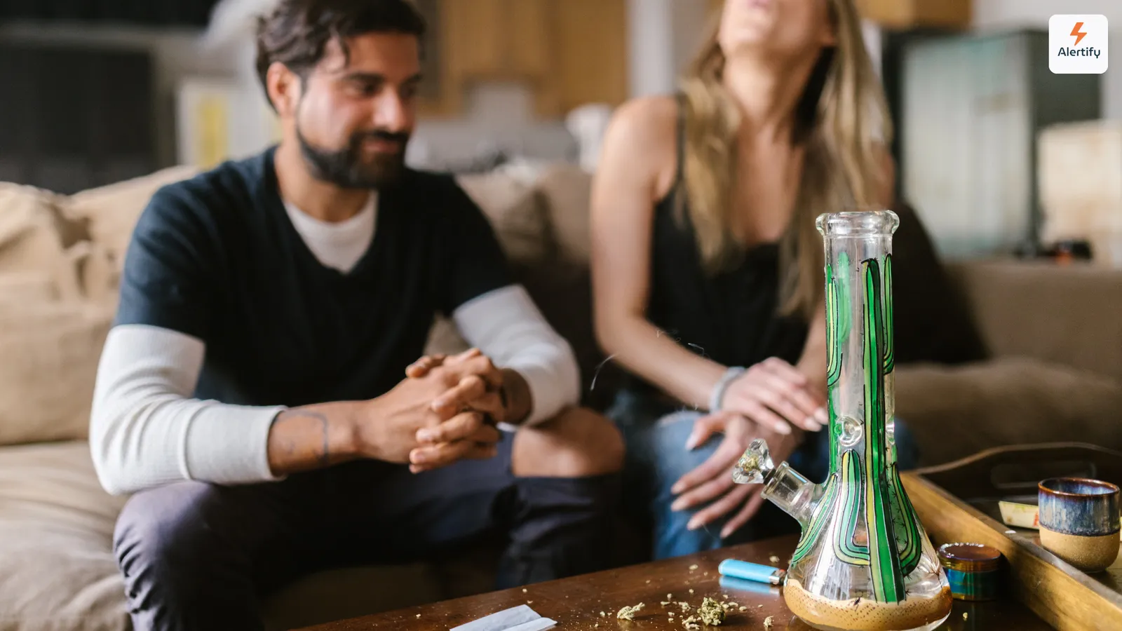 Doi oaspeți așezați pe o canapea în camera de zi a unei închirieri, cu un bong de sticlă, o brichetă și un grinder pe măsuța de cafea din fața lor.